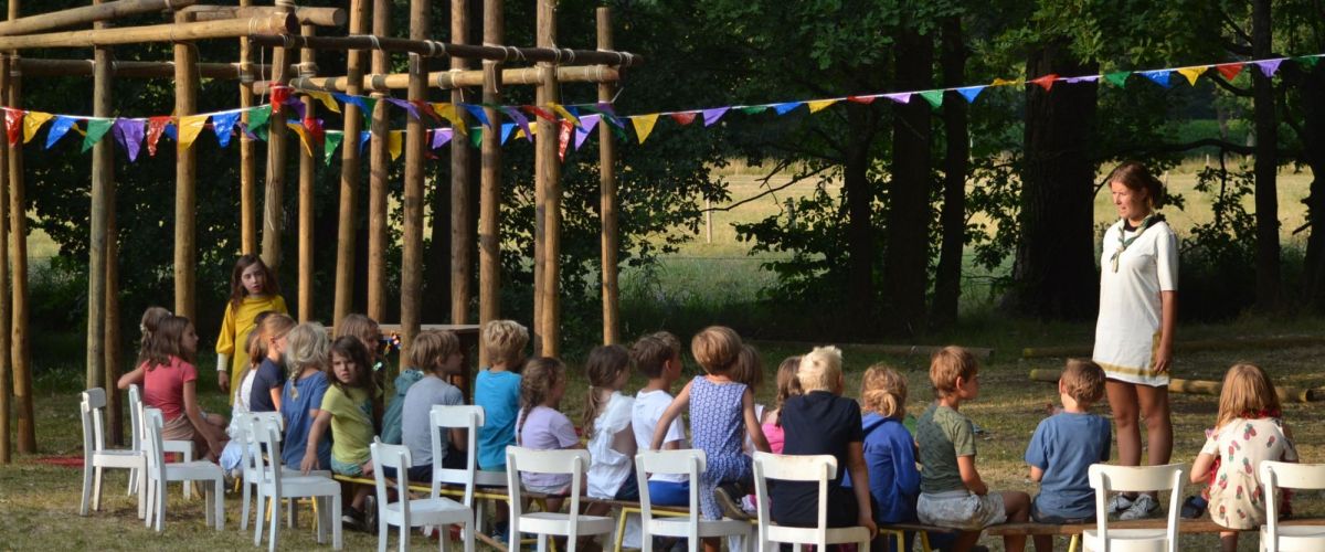 kapoenen op stoeltjes op een grasveld. Naast de kinderen staat een sjorconstructie versiert met vlaggetjes.