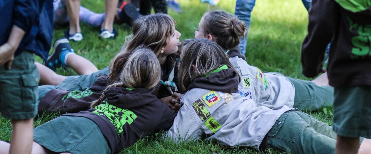 Vier leden in uniform liggen, met hun armen in elkaar gehaakt, in het gras te spelen.