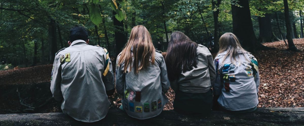 Vier jonge mensen in scoutsuniform die op een omgevallen boomstam in een bos zitten. De ruggen naar de camera gekeerd.