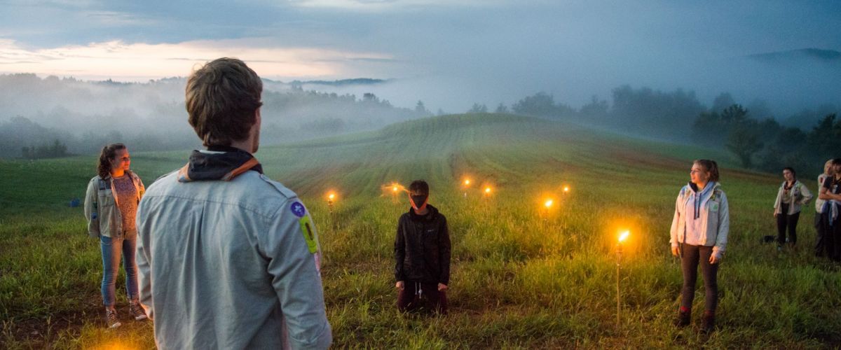 In een glooiend landschap bij zonsopgang zit een geblinddoekte scout op zijn knieën klaar voor zijn totem. Een leider spreekt hem toe.