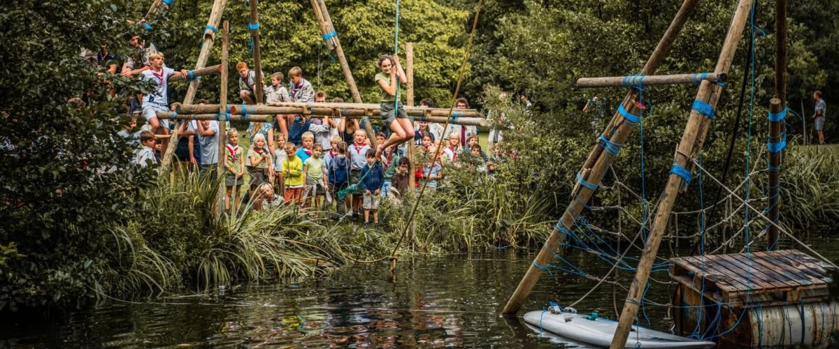 een grote sjorconstructie over een rivier met vanboven een touw waar iemand aan hangt die over de rivier zwiert.