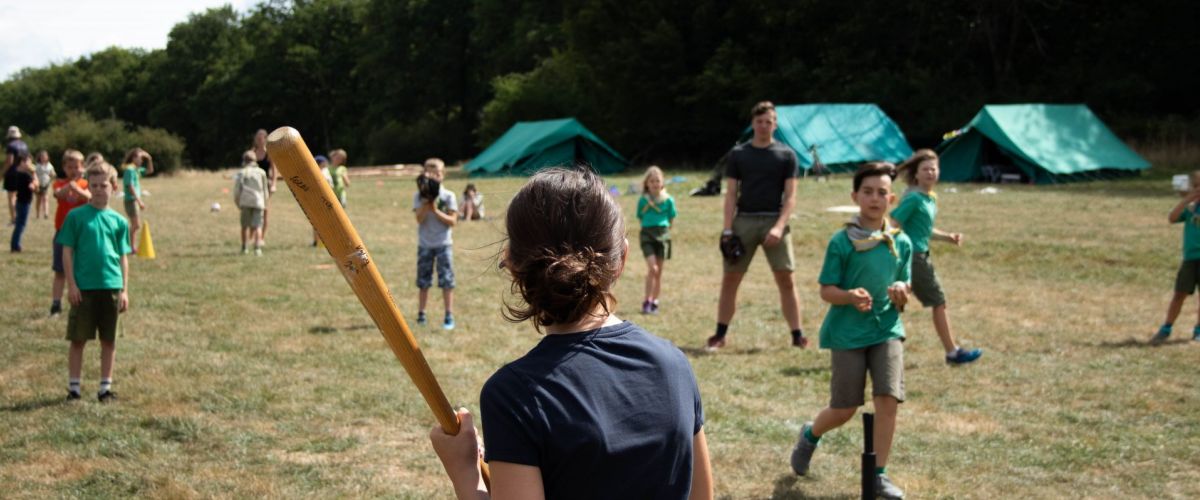 Een meisje staat klaar met een baseballbat, in het veld staan andere spelers klaar. 