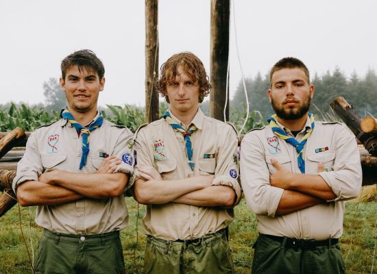 drie scoutsleiders staan op een rijtje, armen over elkaar, op kamp